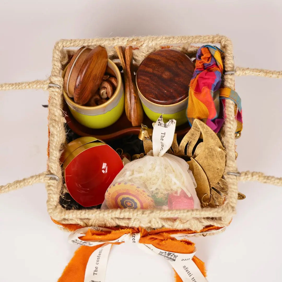 Festive Joy Gift Hamper with Jute Basket, Brass Diyas, Ceramic Tumblers with Dry Fruits, Metal Windchime & Seed Crackers The confetti tree