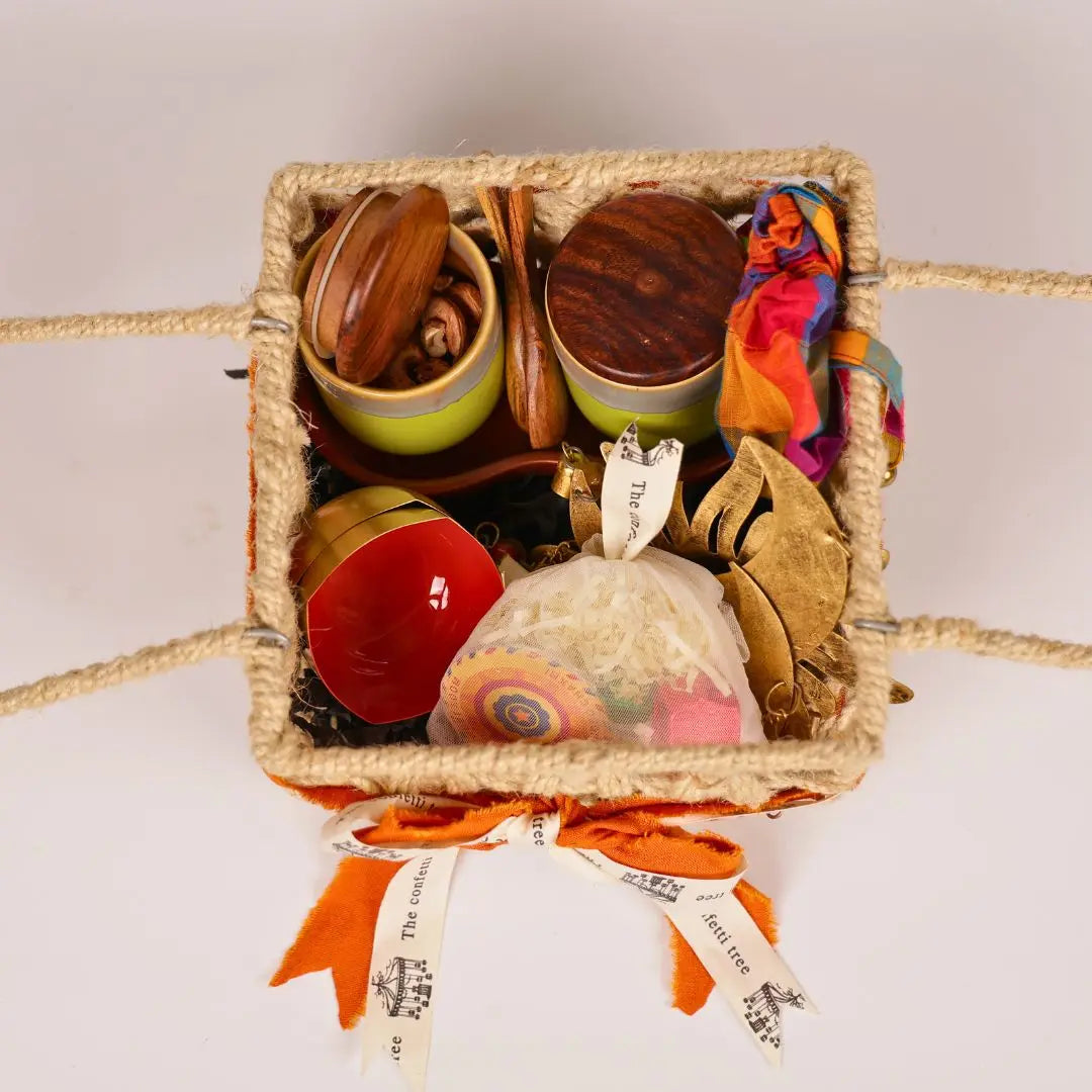 Festive Joy Gift Hamper with Jute Basket, Brass Diyas, Ceramic Tumblers with Dry Fruits, Metal Windchime & Seed Crackers The confetti tree